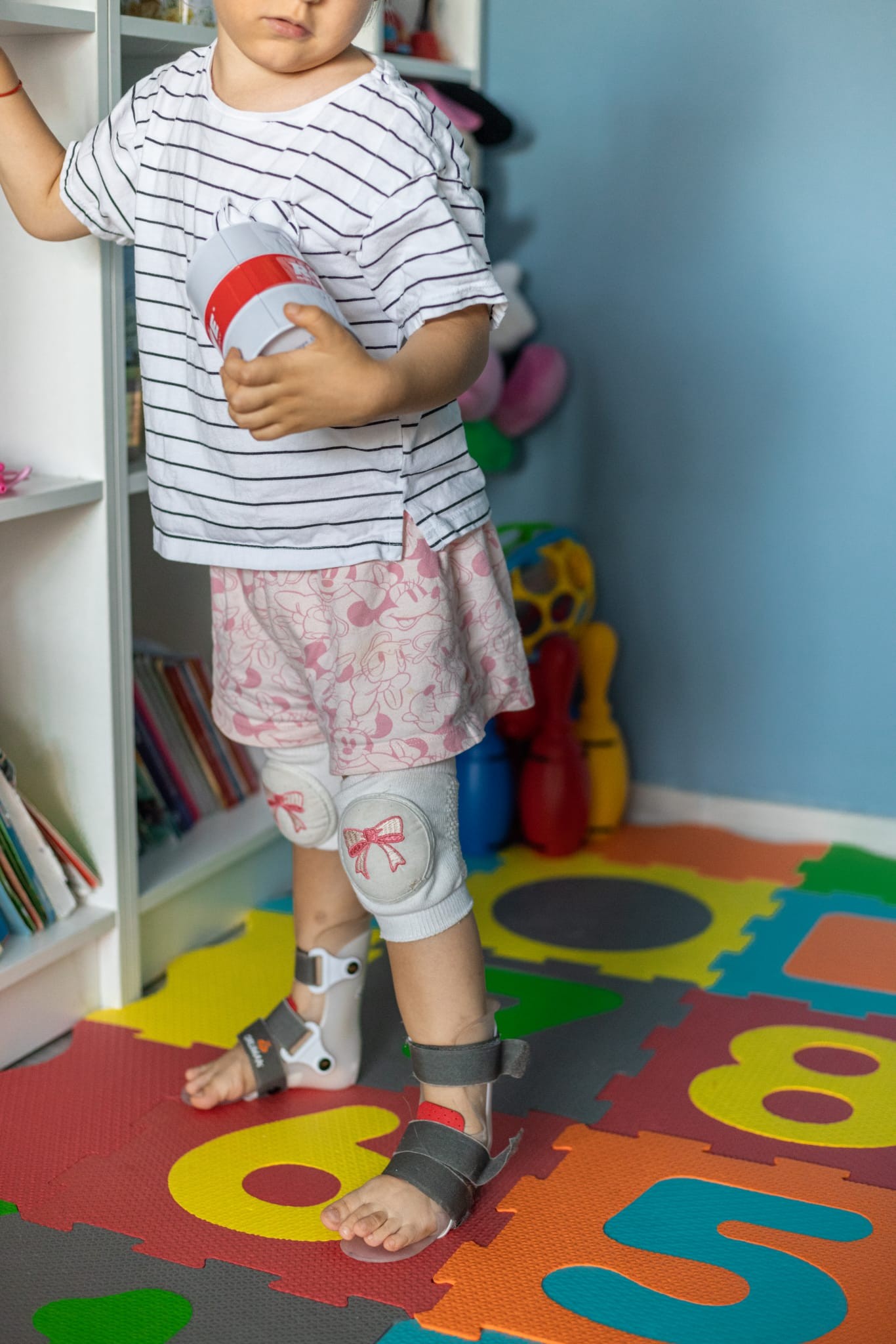 Copil mic stând în picioare purtând orteze la glezne și genunchiere, în cameră de joacă